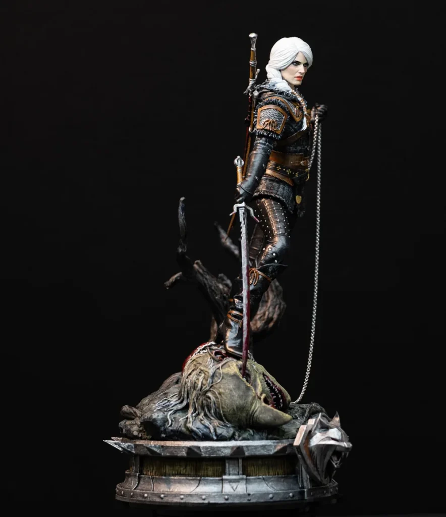 Estátua de Ciri de corpo inteiro, cabelo branco preso, armadura escura, espada apoiada no chão e o pé sobre a cabeça de um monstro